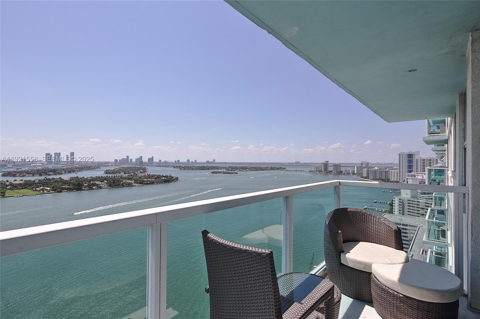 A high-rise balcony features wicker furniture overlooking a bay with a distant city skyline — 650 West Ave # 2801, Miami Beach FL 33139, MIAMI BEACH, FL