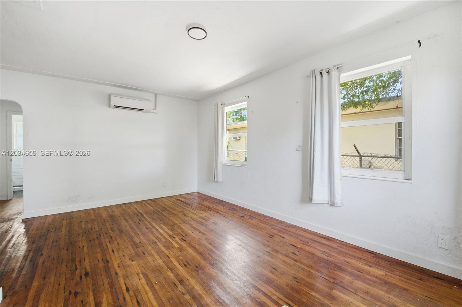 An empty room features hardwood floors, white walls, two windows, and a wall-mounted air conditioner — 101 NW 74th St # 4, Miami FL 33150, MIAMI, FL