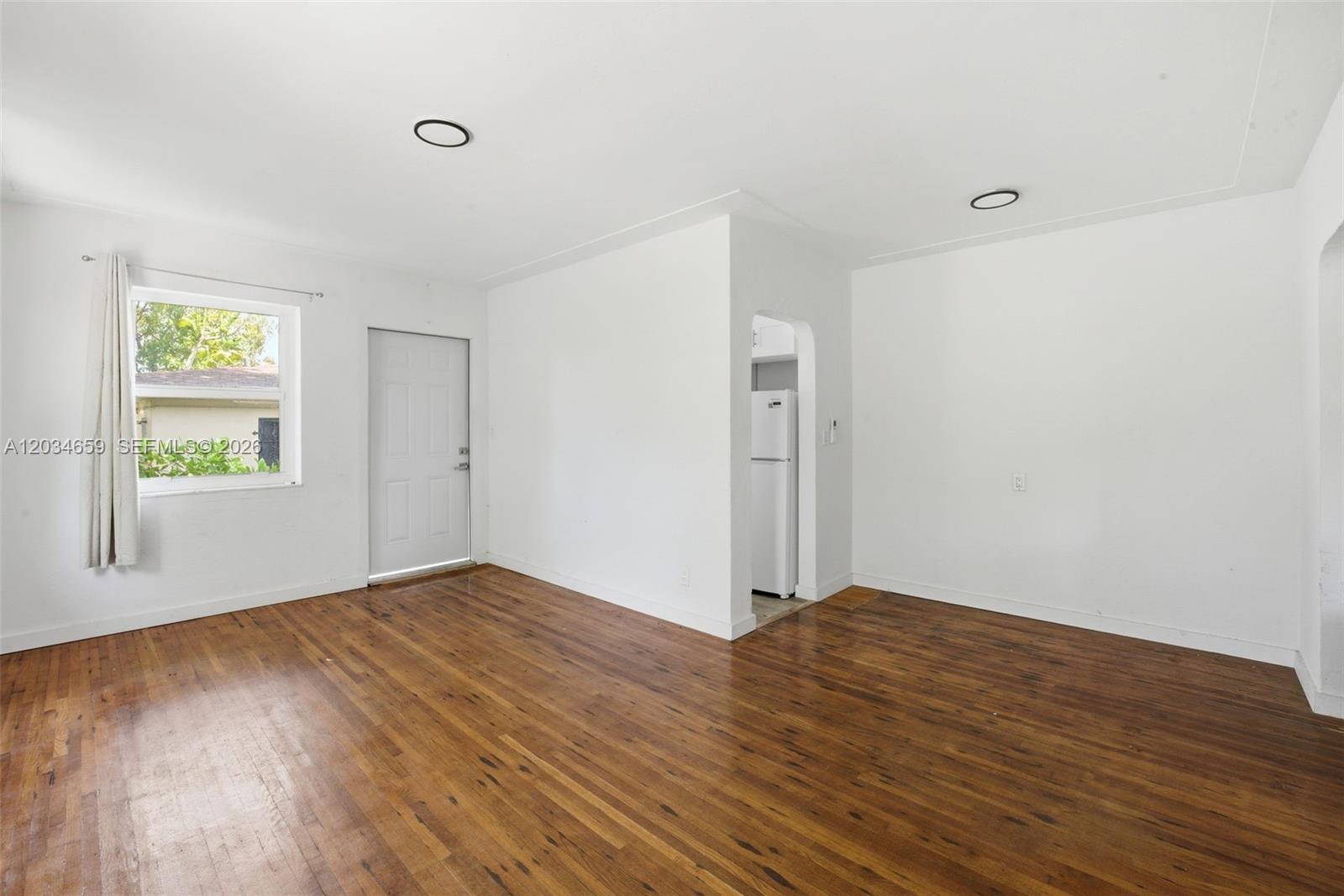 An empty room features white walls, hardwood floors, a window, and a doorway to a kitchen — 101 NW 74th St # 4, Miami FL 33150, MIAMI, FL