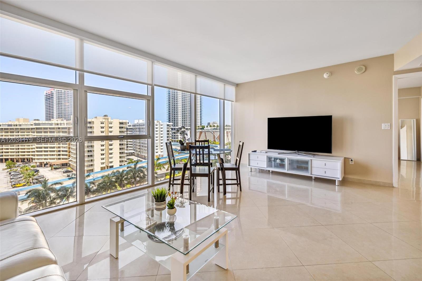 A living room features large floor-to-ceiling windows, tile flooring, and a view of city buildings — 1830 S Ocean Dr # 907, Hallandale Beach FL 33009, HALLANDALE BEACH, FL