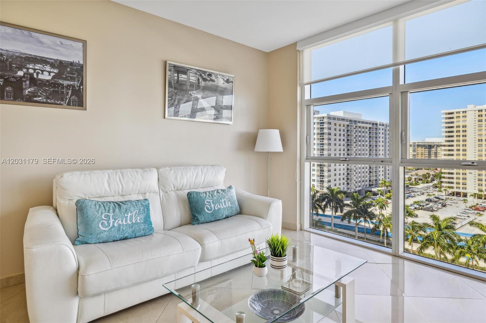 A living room features a white sofa, glass coffee table, and large windows overlooking buildings — 1830 S Ocean Dr # 907, Hallandale Beach FL 33009, HALLANDALE BEACH, FL