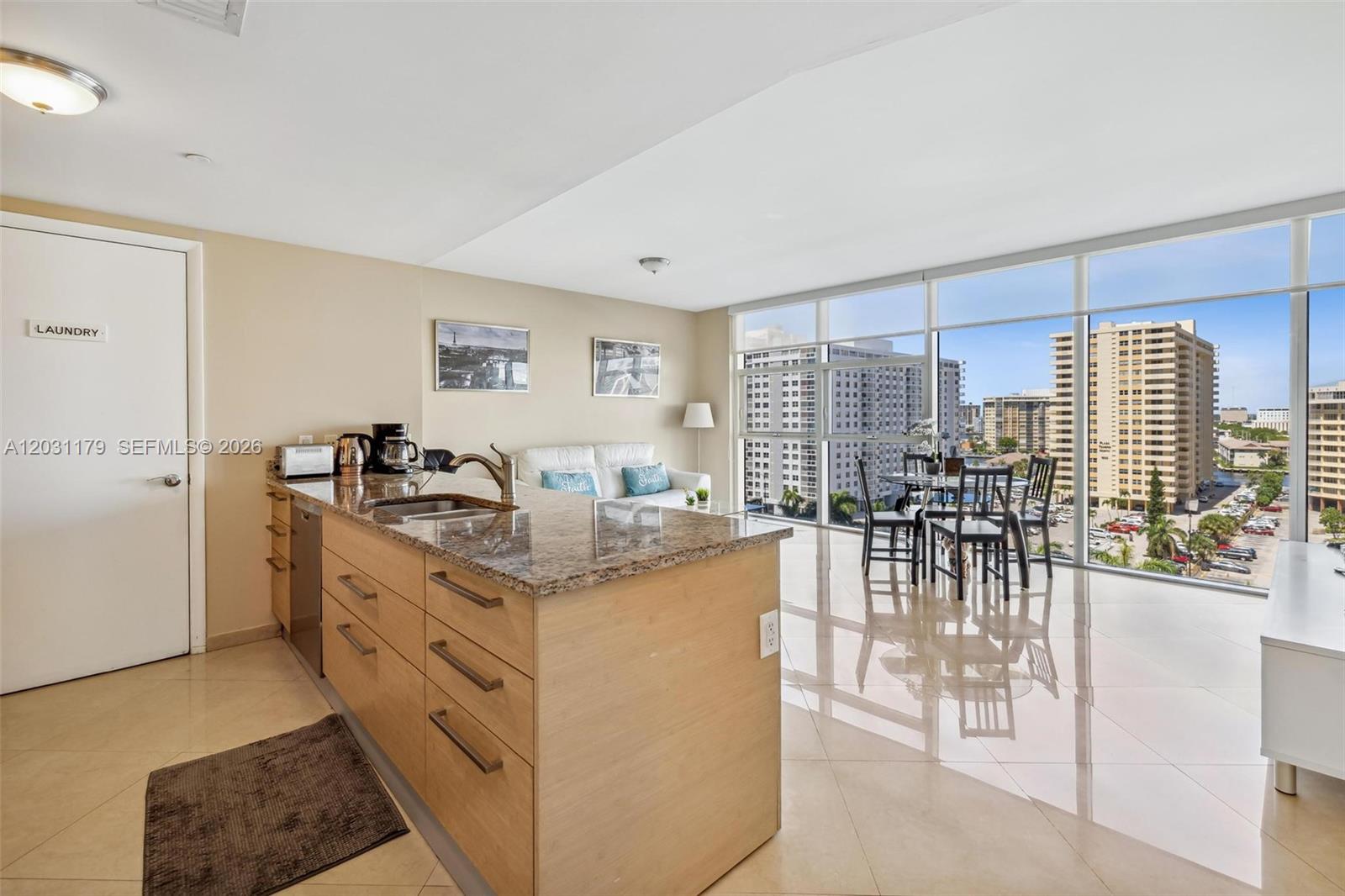 A modern kitchen with granite countertops and light wood cabinets overlooks a bright living area — 1830 S Ocean Dr # 907, Hallandale Beach FL 33009, HALLANDALE BEACH, FL