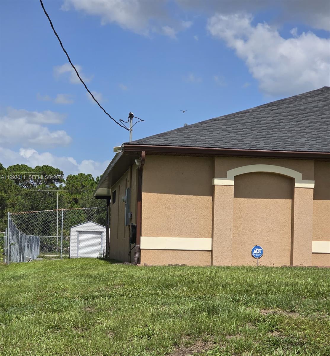 The exterior of a tan stucco house features a dark shingled roof and a chain-link fence — 1816 Tomaso Ave, Lehigh Acres FL 33972, LEHIGH ACRES, FL