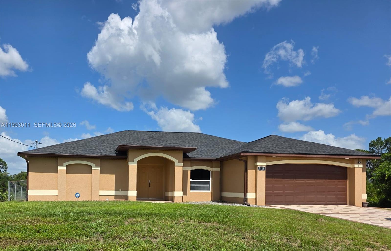 A single-story tan house with a dark shingled roof and a two-car garage — 1816 Tomaso Ave, Lehigh Acres FL 33972, LEHIGH ACRES, FL