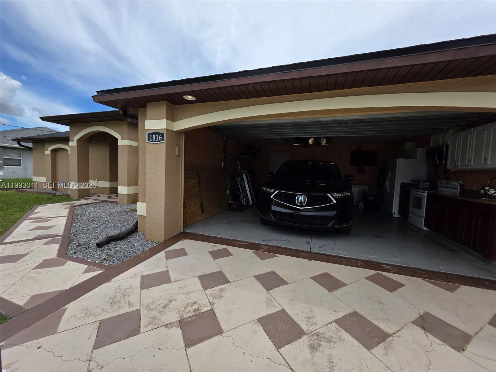 A tan house exterior features a paved driveway and a garage containing a black car — 1816 Tomaso Ave, Lehigh Acres FL 33972, LEHIGH ACRES, FL