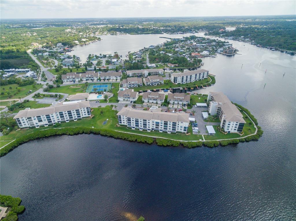 An aerial view shows waterfront condominium buildings surrounded by trees, a tennis court, and a pool — 8211 BRENT STREET UNIT 816, PORT RICHEY, FL