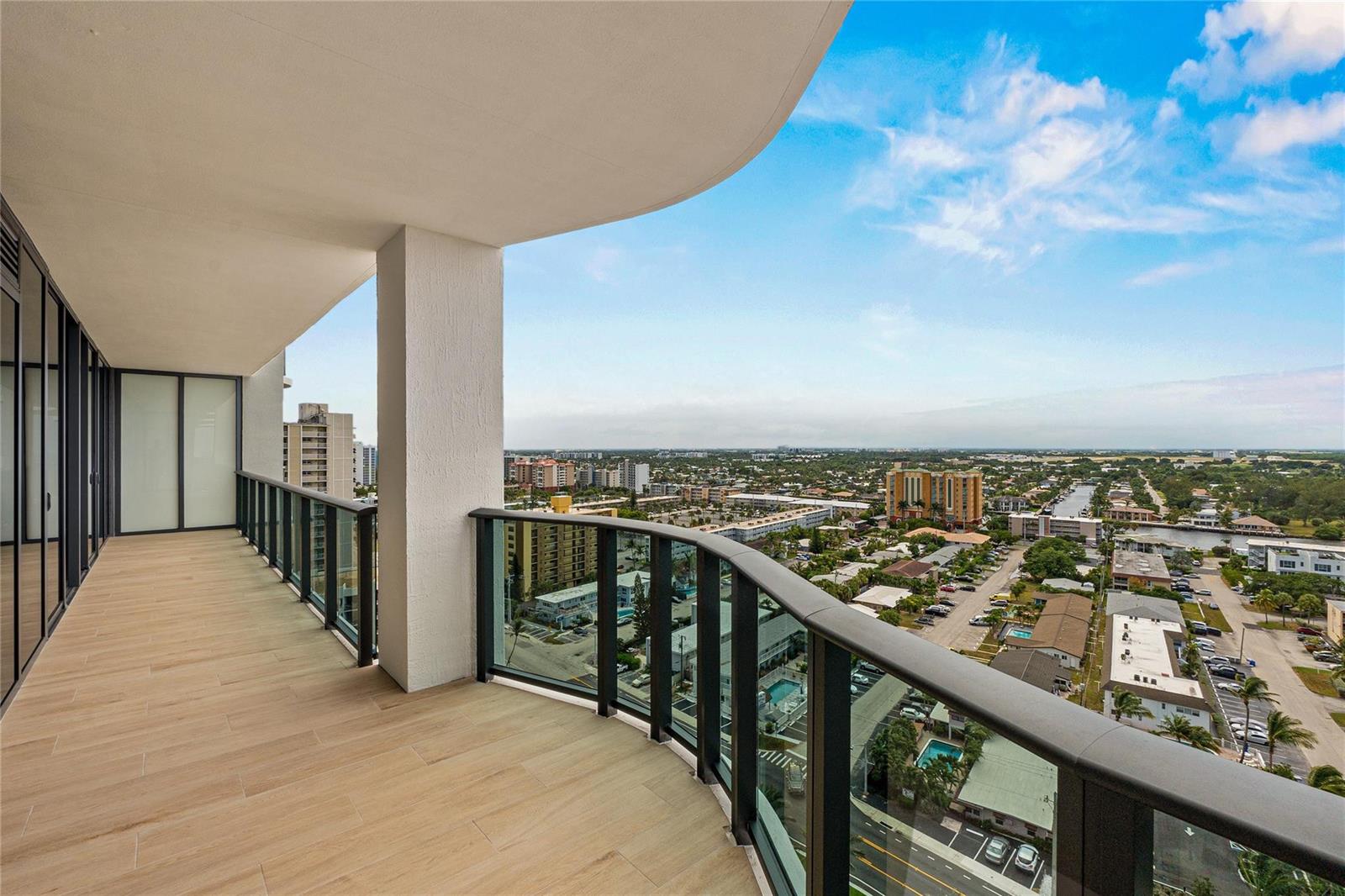 A high-rise balcony with wood-look tile flooring and a glass railing overlooking a suburban landscape — 900 N Ocean Boulevard # 1604, Pompano Beach FL 33062, POMPANO BEACH, FL