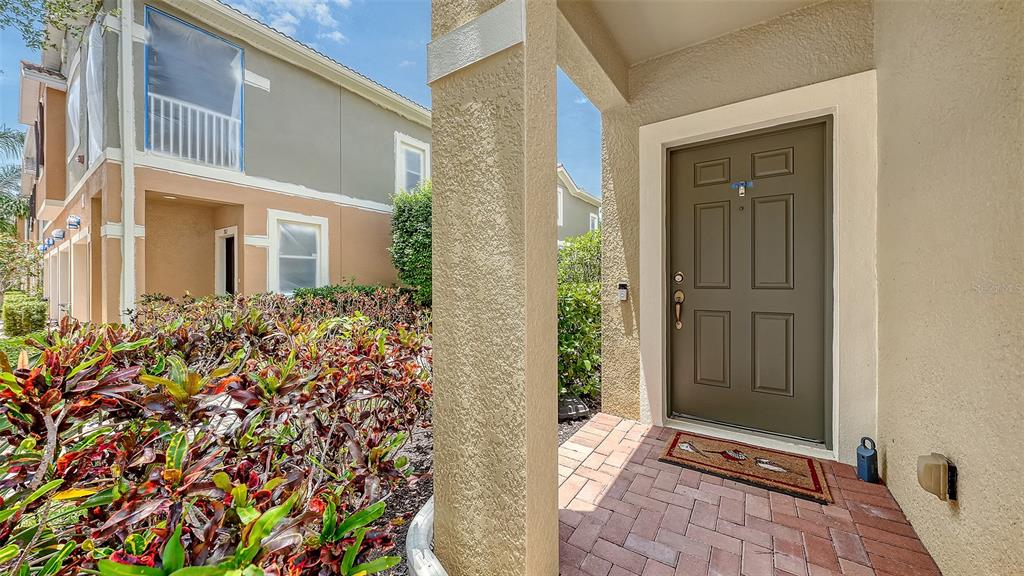 A brown front door with a brick porch and colorful landscaping in front of a building — 7843 MOONSTONE DRIVE UNIT 24-202, SARASOTA, FL