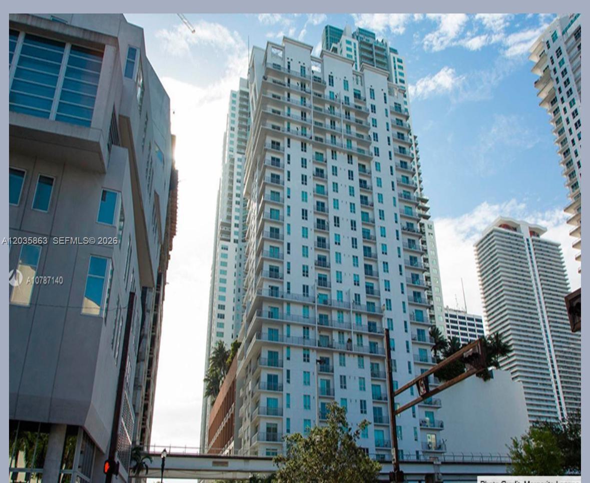 A tall white residential high-rise building stands between other urban structures under a blue sky — 234 NE 3rd St # 1506, Miami FL 33132, MIAMI, FL