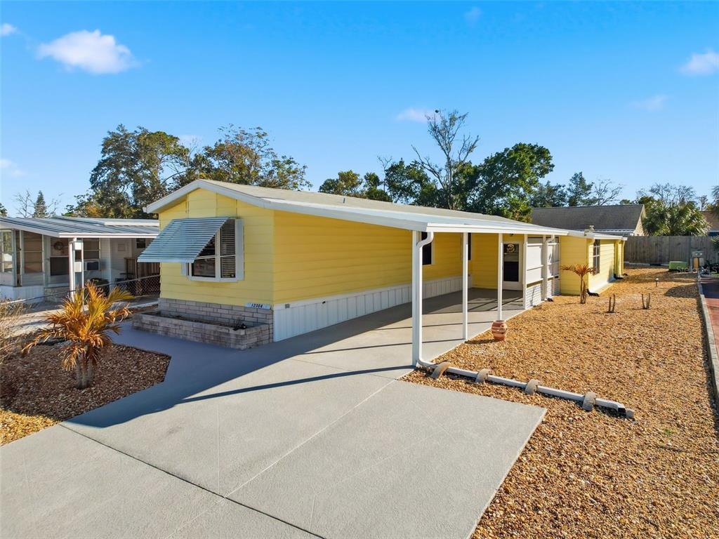 A yellow mobile home with a concrete driveway, carport, and gravel landscaping under a blue sky — 12304 LONGHORN DRIVE, HUDSON, FL