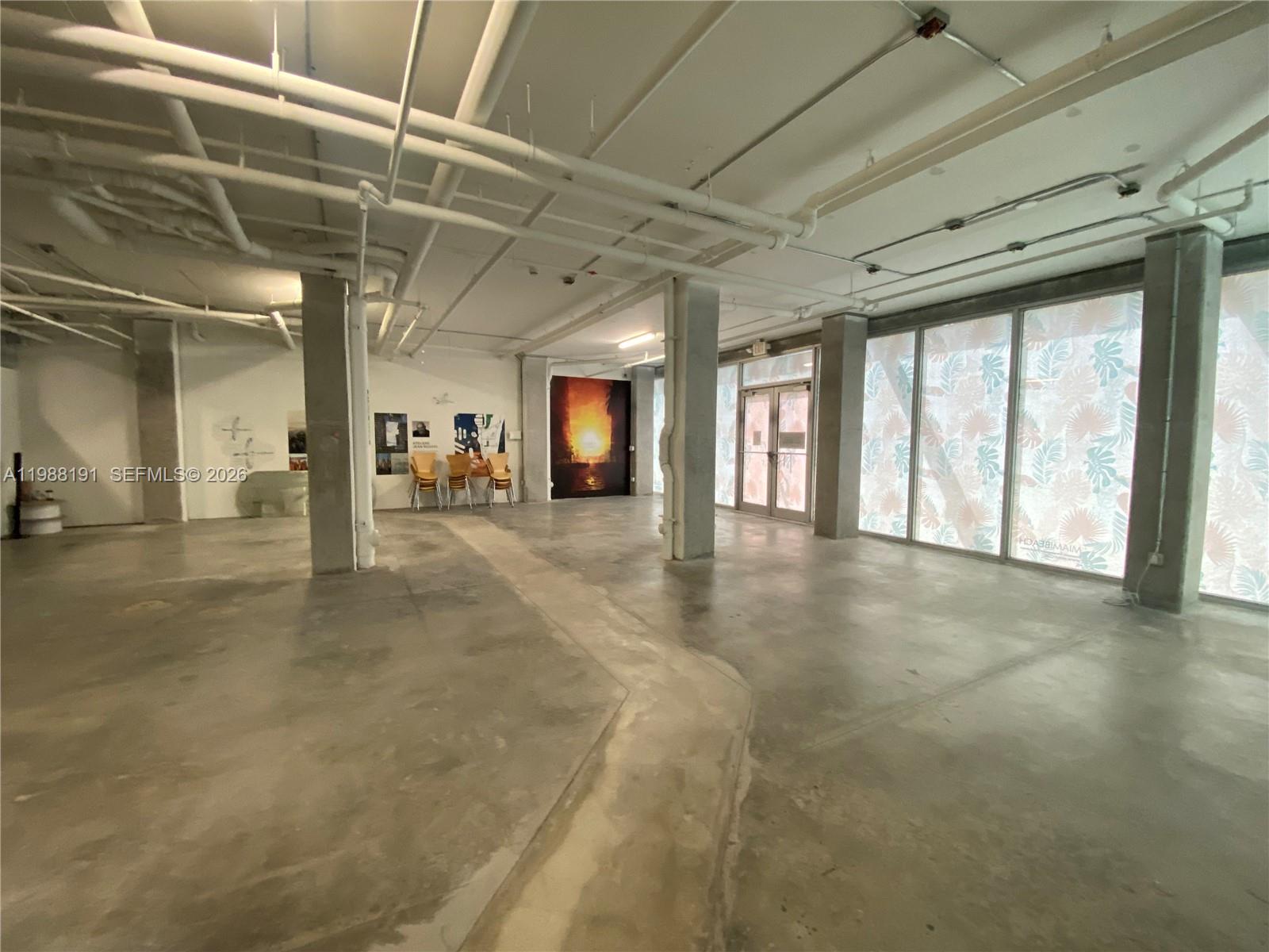 An open-concept commercial space features exposed ceiling pipes, concrete floors, and large glass windows — 1215 West Ave # 1, Miami Beach FL 33139, MIAMI BEACH, FL