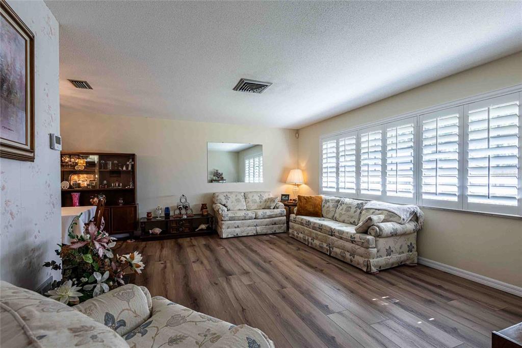 A living room features wood-look flooring, floral-patterned sofas, and white plantation shutters on the windows — 1830 OAK FOREST DRIVE S, CLEARWATER, FL
