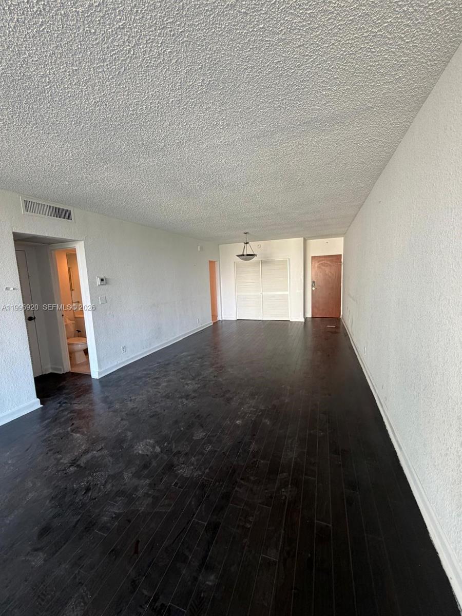 An empty room features dark wood flooring, white walls, a textured ceiling, and a bathroom entrance — 4001 S Ocean Dr # 4H, Hollywood FL 33019, HOLLYWOOD, FL