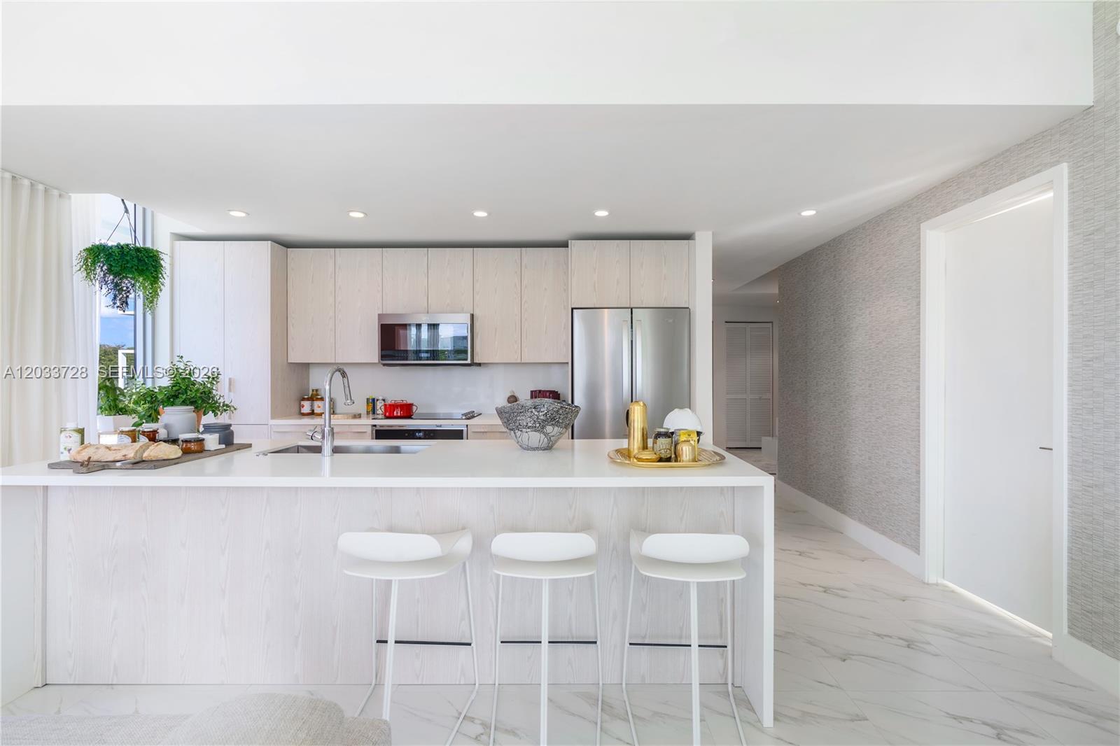 A modern kitchen features light wood cabinetry, a white island with bar stools, and marble flooring — 2000 Metropica Way # 203, Sunrise FL 33323, SUNRISE, FL