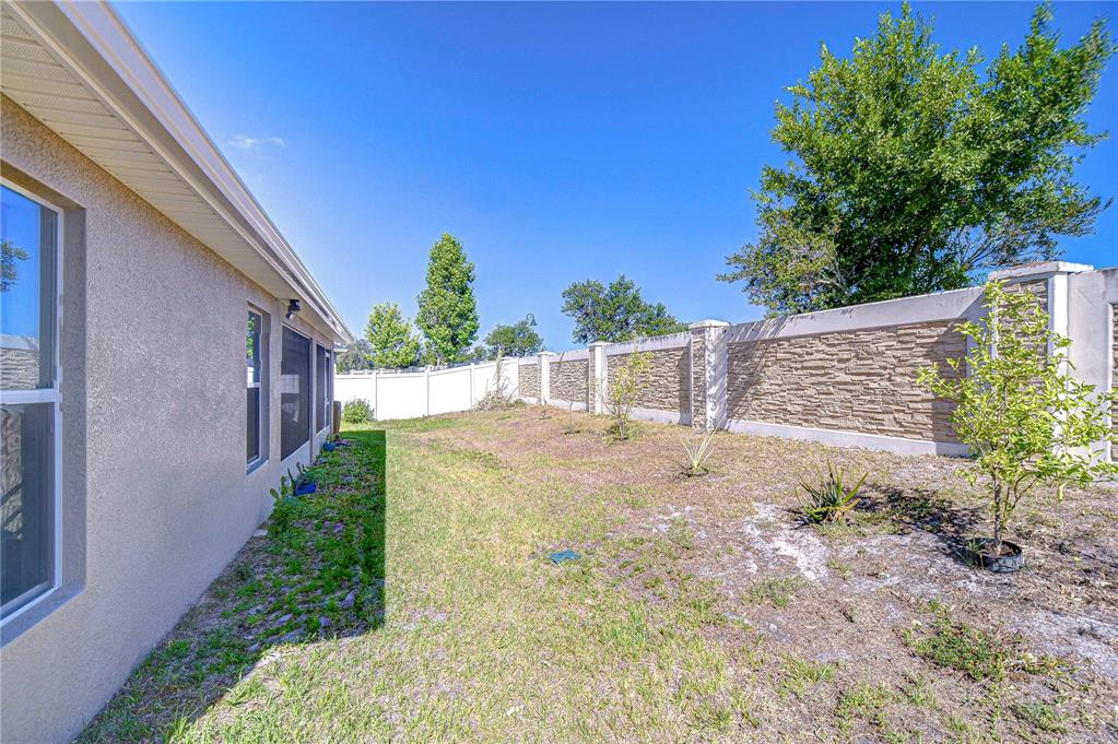 A side yard features a beige house wall, a stone-textured privacy wall, and sparse landscaping — 6731 CASTLE GREEN PLACE, ZEPHYRHILLS, FL