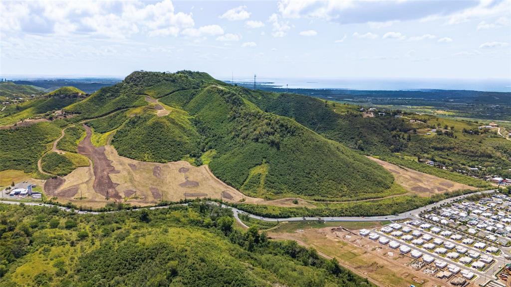 An aerial view shows a large green mountain rising above a residential neighborhood and road — PR-153 KM 10.7 BO. RIO JUEYES, SANTA ISABEL, PR
