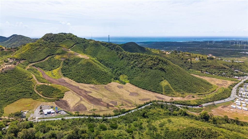 An aerial view shows a large, hilly landscape with cleared land and a nearby road — PR-153 KM 10.7 BO. RIO JUEYES, SANTA ISABEL, PR