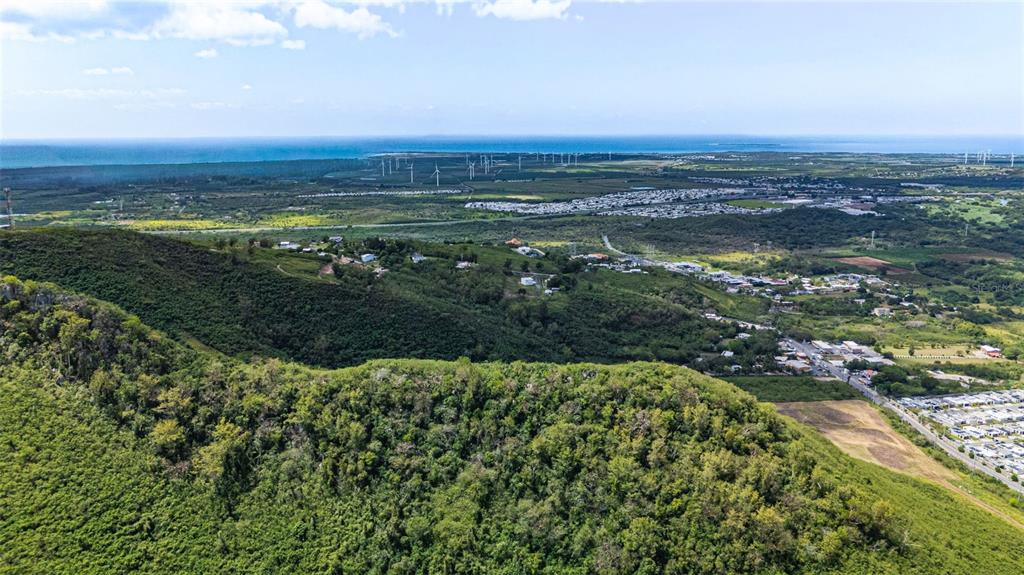 An aerial view shows a lush, hilly landscape overlooking a coastal plain with wind turbines — PR-153 KM 10.7 BO. RIO JUEYES, SANTA ISABEL, PR