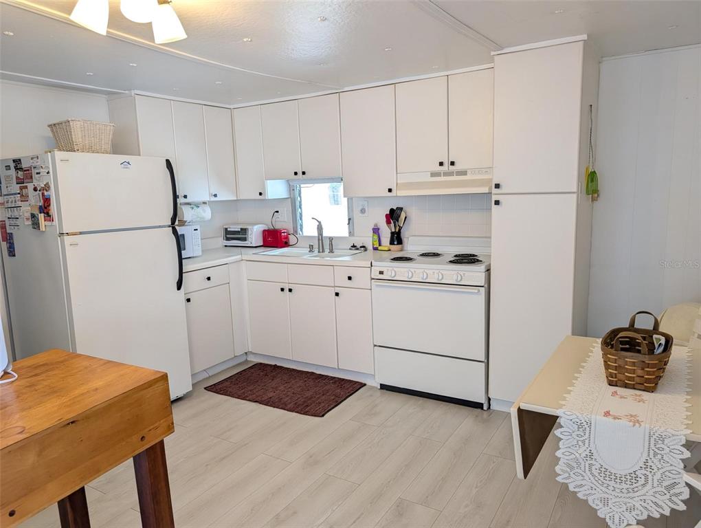 A kitchen features white cabinets, white appliances, light wood-look flooring, and a small dining table — 1702 OLYMPIA FIELDS STREET, SARASOTA, FL