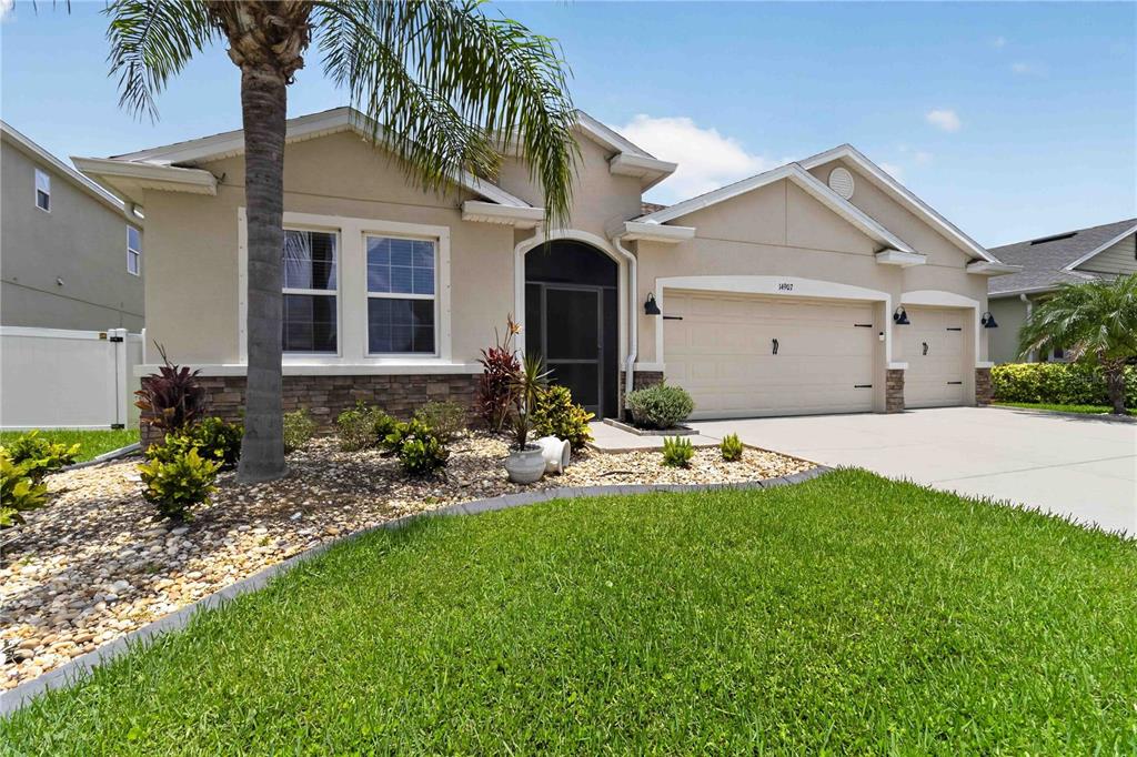 A single-story suburban house with a tan stucco exterior, stone accents, and a three-car garage — 14907 TRINITY FALL WAY, BRADENTON, FL