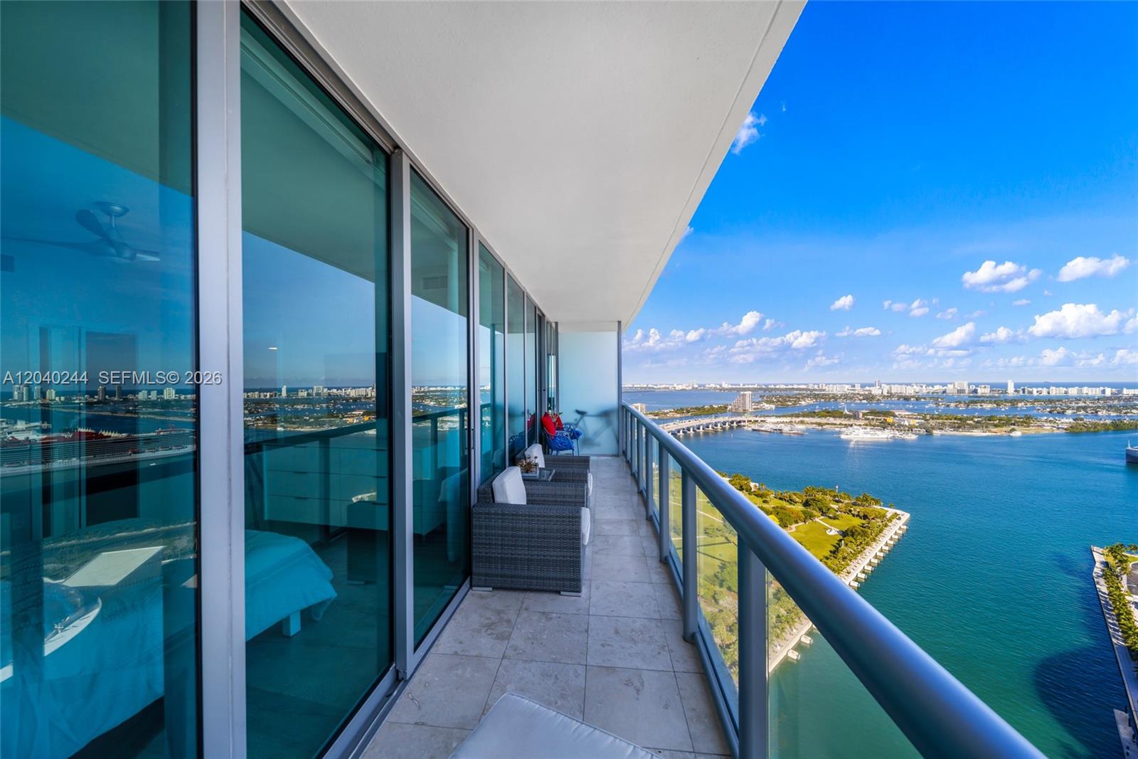 A high-rise balcony features gray wicker furniture, glass railings, and a panoramic waterfront view — 888 Biscayne Blvd # 3907, Miami FL 33132, MIAMI, FL