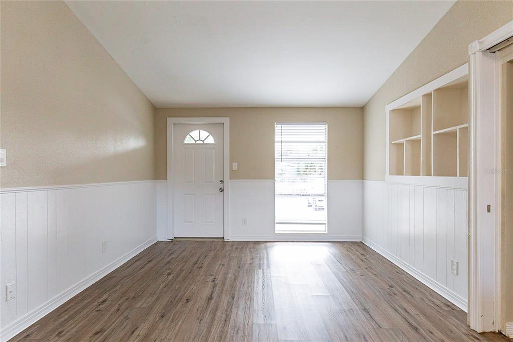 An empty room features wood-look flooring, white wainscoting, a front door, and built-in wall shelving — 7216 E EMMA STREET, TAMPA, FL