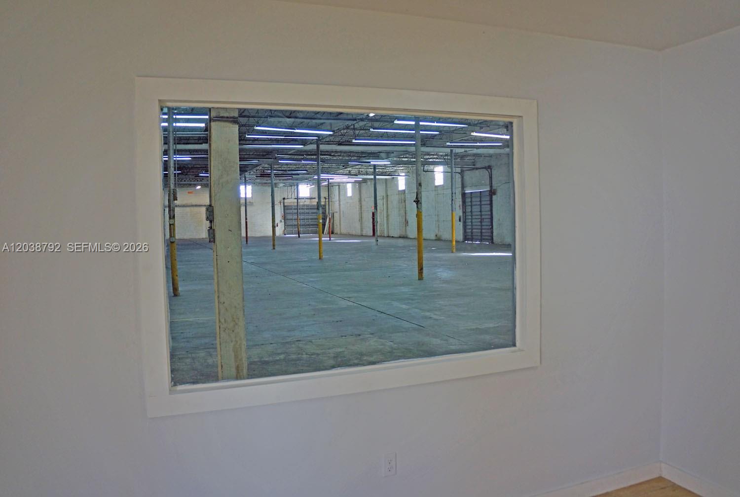 A white-walled room features a large rectangular window looking into a spacious industrial warehouse — 3690 NW 52nd St, Hialeah FL 33142, HIALEAH, FL