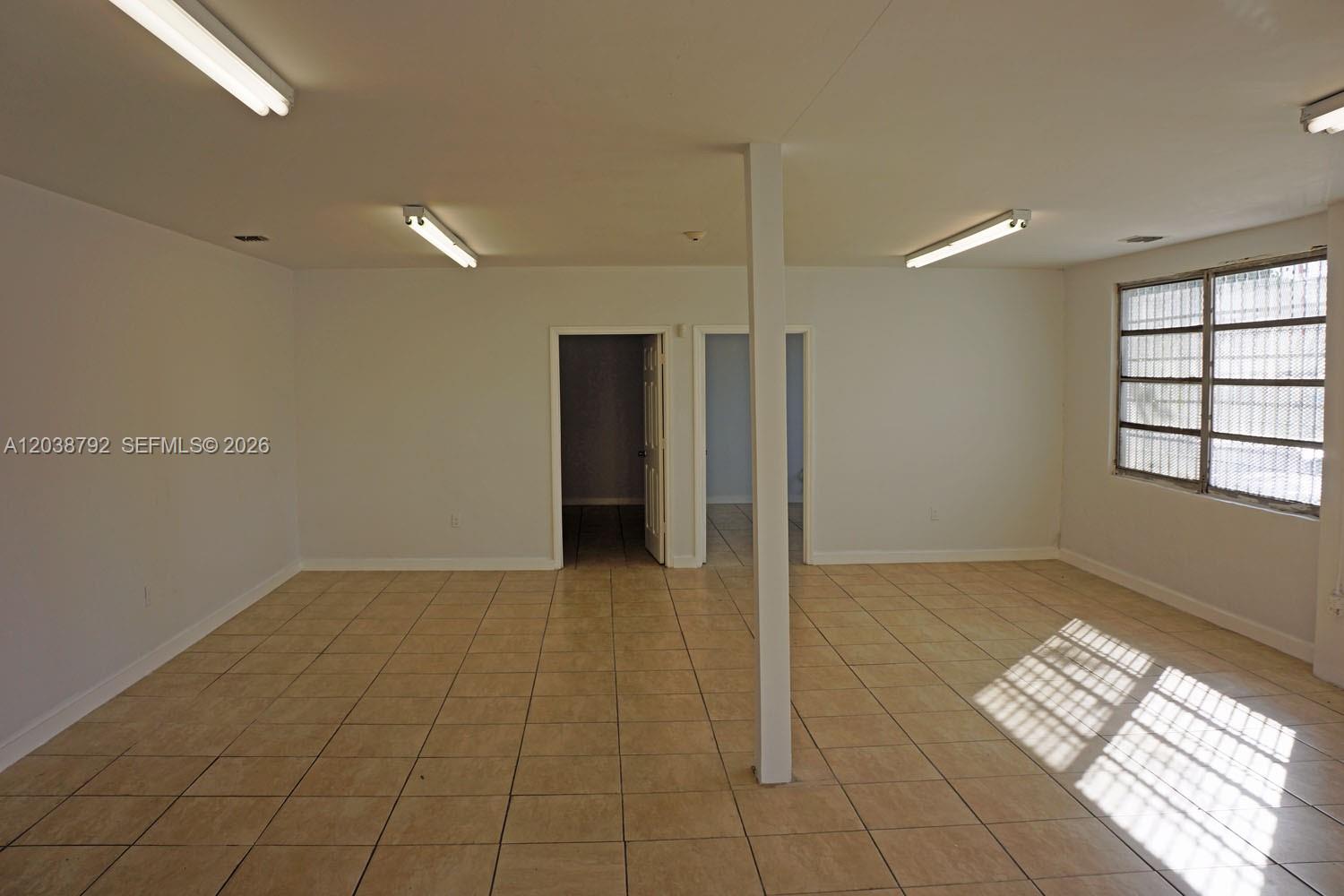 An empty room features beige tile flooring, white walls, a central support column, and fluorescent lighting — 3690 NW 52nd St, Hialeah FL 33142, HIALEAH, FL