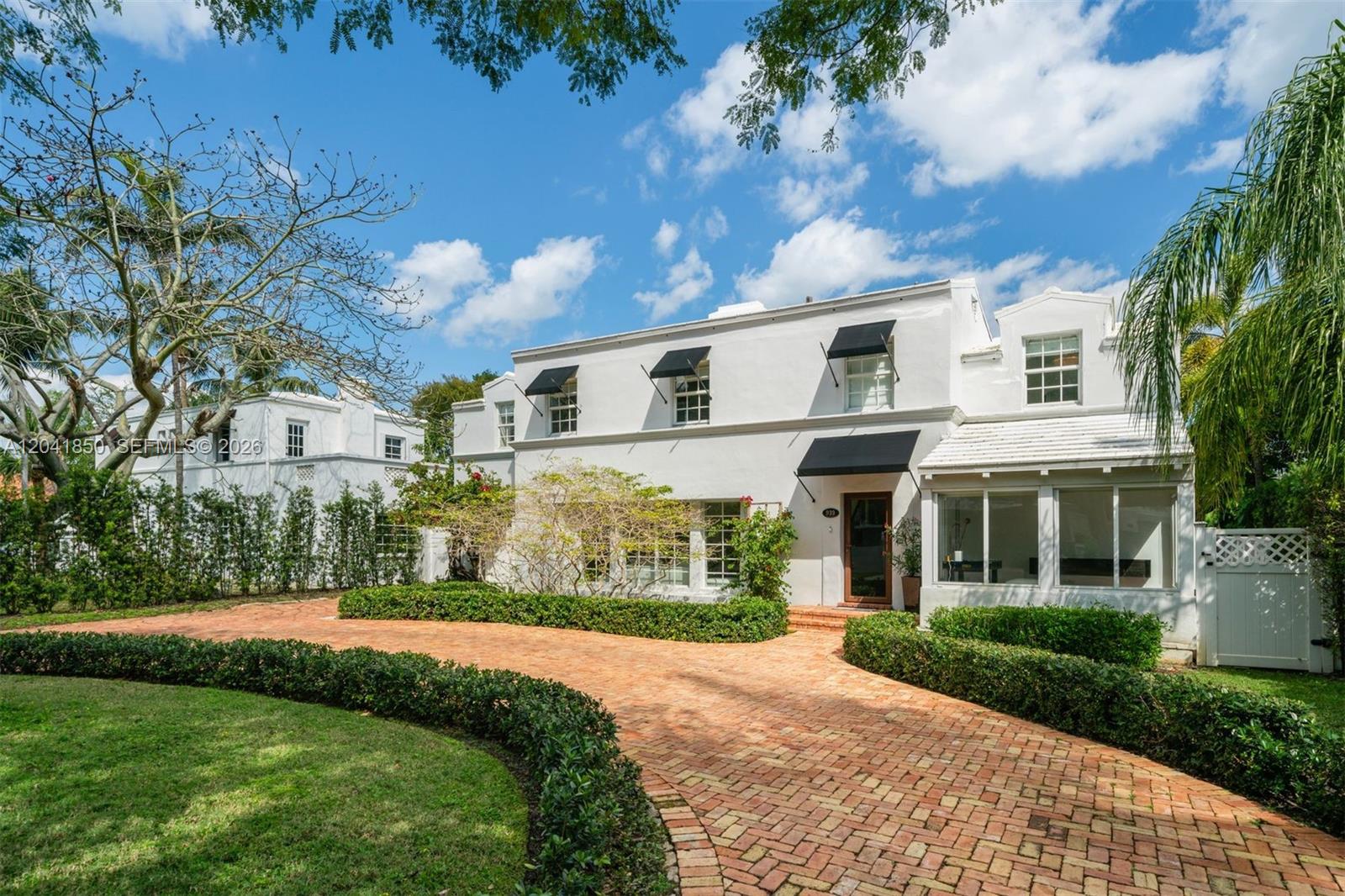 A two-story white house features a brick driveway, black awnings, and manicured hedges — 939 W 47th Ct, Miami Beach FL 33140, MIAMI BEACH, FL