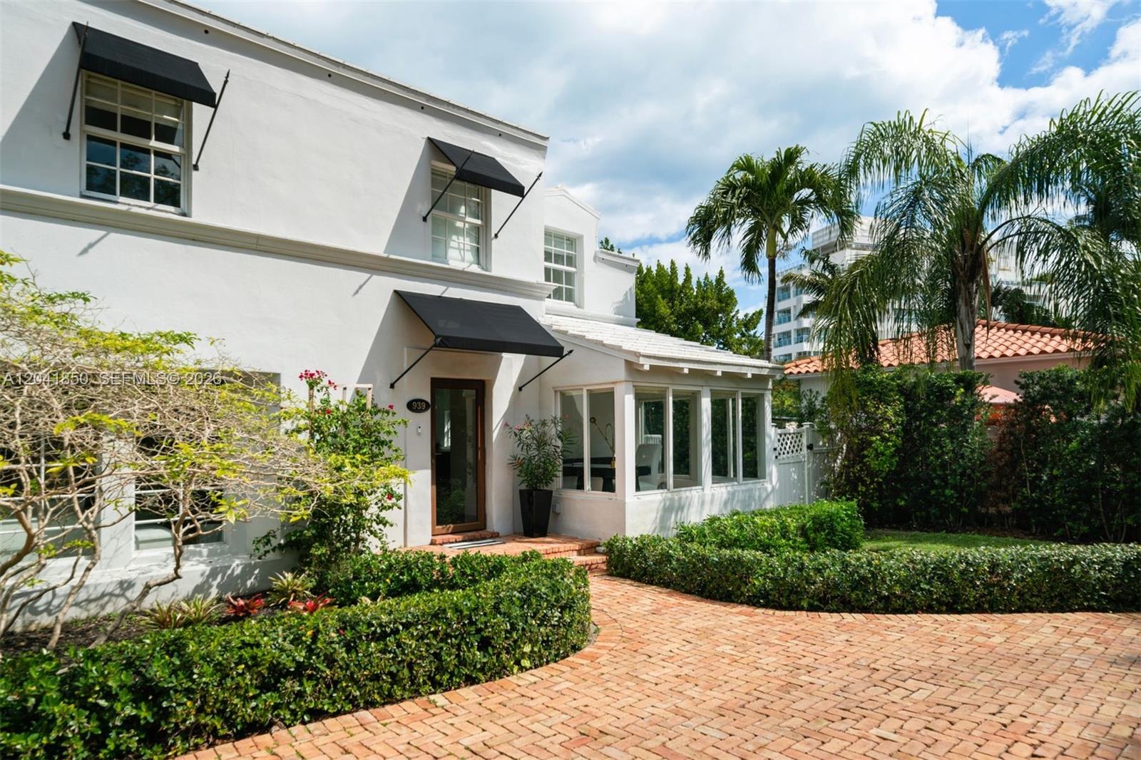 A white two-story house features black window awnings, a brick driveway, and lush landscaping — 939 W 47th Ct, Miami Beach FL 33140, MIAMI BEACH, FL