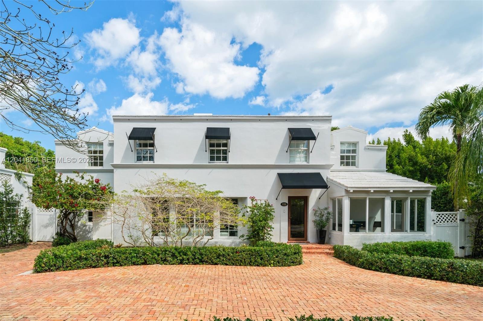 A two-story white house features black awnings, a brick driveway, and lush landscaping — 939 W 47th Ct, Miami Beach FL 33140, MIAMI BEACH, FL