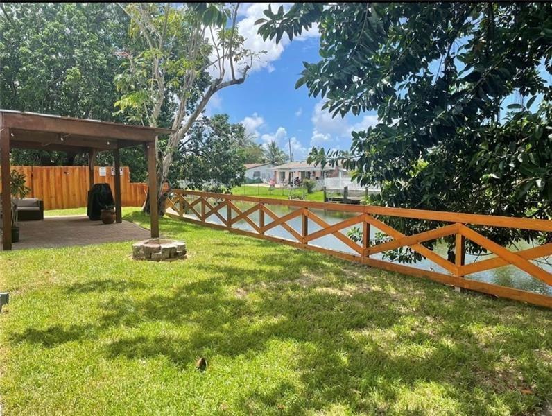 A grassy backyard features a wooden pergola, a fire pit, and a fence bordering a canal — 5620 SW 56th Street, Davie FL 33314, DAVIE, FL