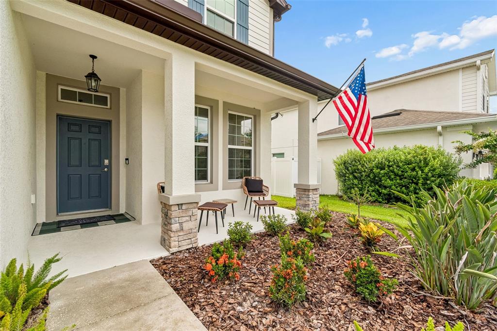 A covered front porch features a blue door, stone-based columns, and a small seating area — 23958 TERRACINA COURT, LAND O LAKES, FL