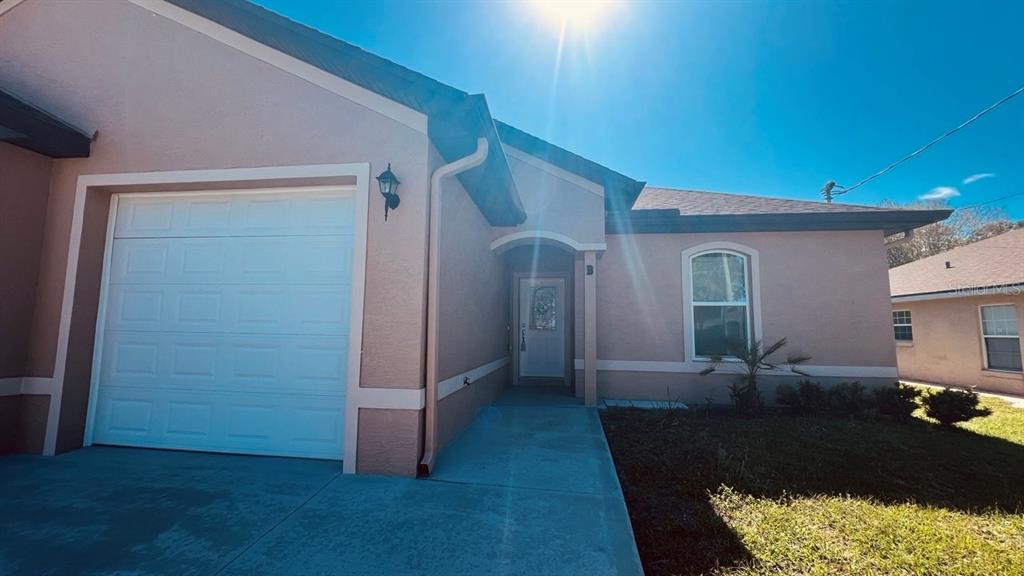 A tan stucco house exterior features a white garage door, a front entrance, and landscaping — 57 COLUMBIA LANE UNIT B, PALM COAST, FL