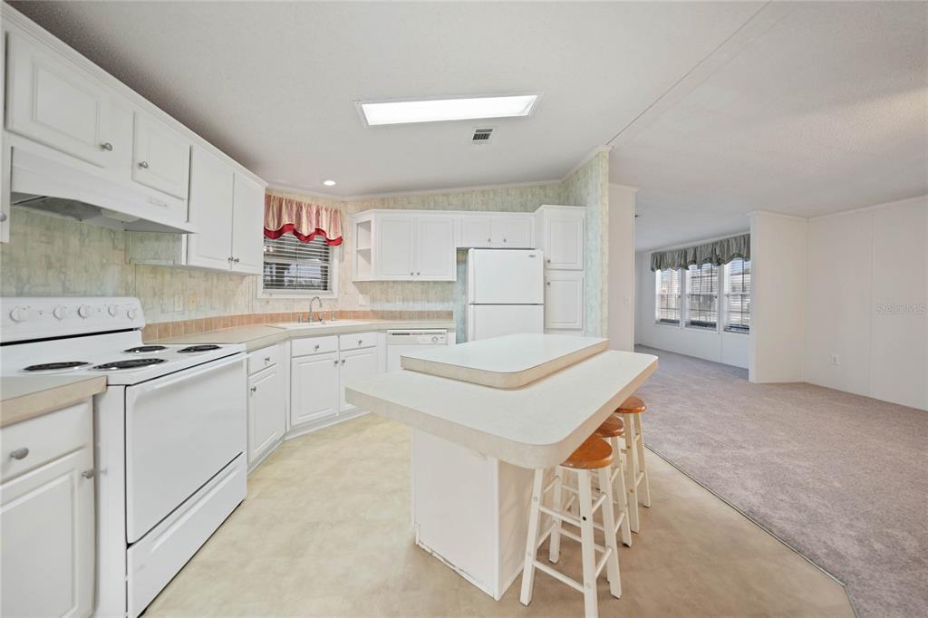 A kitchen with white cabinets and an island sits adjacent to a carpeted living area — 2100 KINGS HIGHWAY UNIT 517, PORT CHARLOTTE, FL