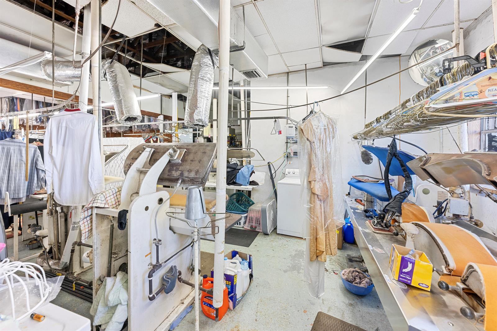 A commercial laundry room features industrial pressing equipment, hanging garments, and a white washing machine — 400 S Parrott Avenue, Okeechobee FL 34974, OKEECHOBEE, FL