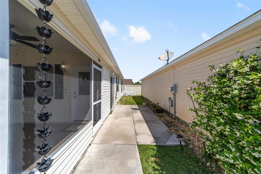 A narrow concrete walkway sits between two houses with a rain chain in the foreground — 2403 BATON PLACE, THE VILLAGES, FL