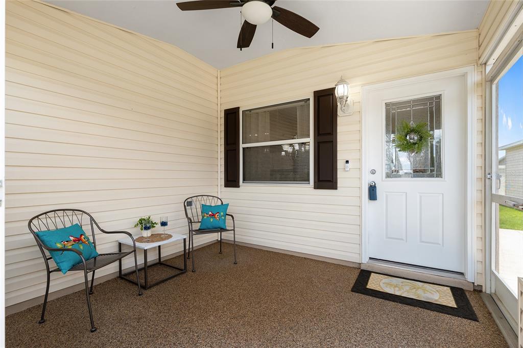 A covered porch with beige siding, a ceiling fan, two chairs, and a white door — 2403 BATON PLACE, THE VILLAGES, FL