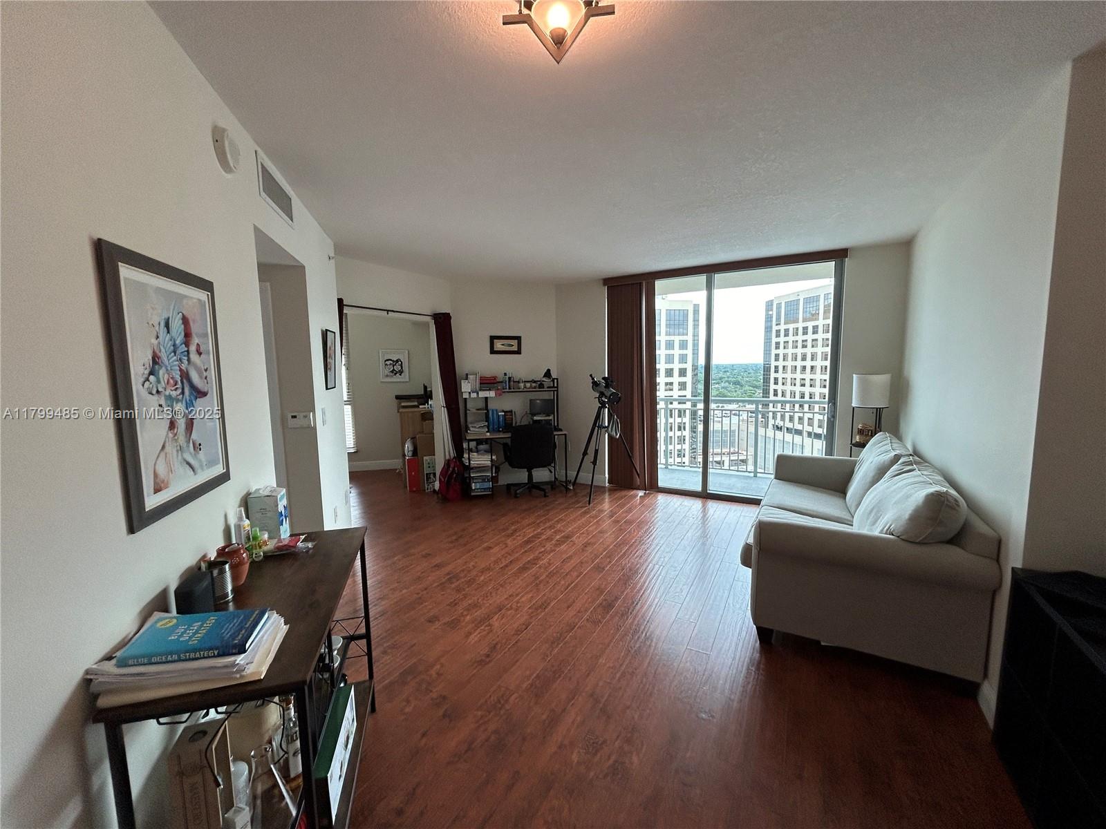A living room with wood-look flooring, a sofa, a desk, and a balcony with city views — 9066 SW 73rd Ct # 1504, Miami FL 33156, MIAMI, FL