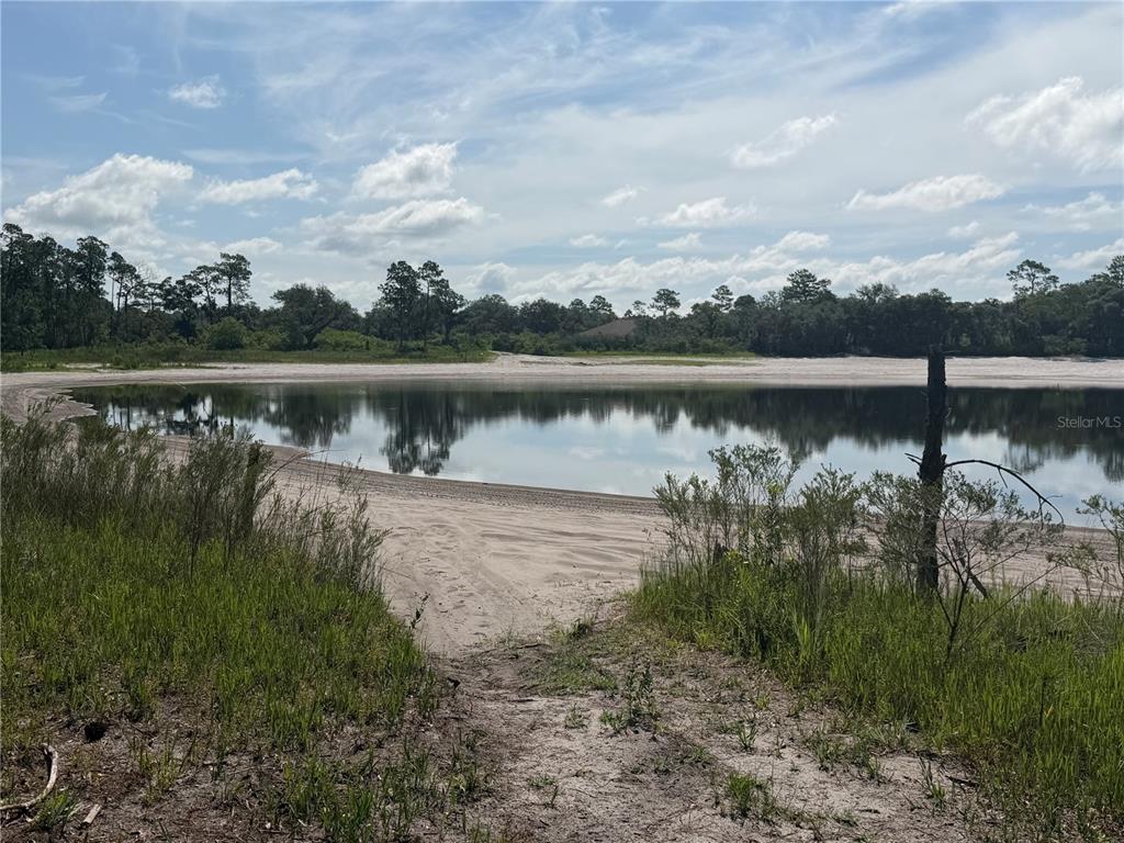 A sandy shoreline borders a calm pond surrounded by trees under a cloudy sky — TBD MALAUKA PASS TRAIL, OCKLAWAHA, FL