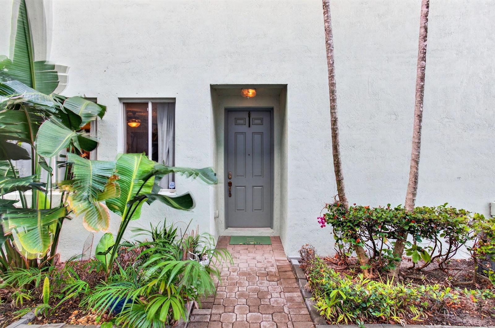 A gray front door is centered on a stucco wall with a brick walkway and landscaping — 3167 Laurel Ridge Circle, Riviera Beach FL 33404, RIVIERA BEACH, FL