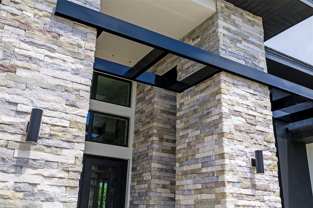 A modern home entrance features stacked stone walls, black metal beams, and a glass door — 4116 BUTTE TRAIL, BRADENTON, FL