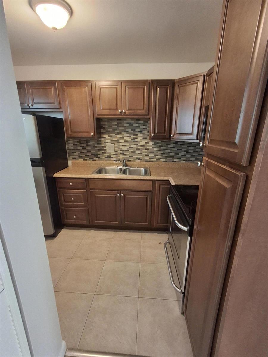 A kitchen features brown wooden cabinets, a mosaic tile backsplash, and stainless steel appliances — 512 Gardens Drive # 103, Pompano Beach FL 33069, POMPANO BEACH, FL