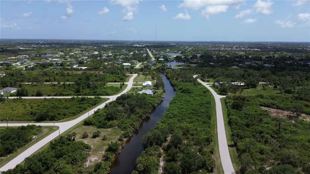An aerial view shows a canal running between two paved roads and surrounding wooded lots — 7711 COSMO COURT, PORT CHARLOTTE, FL