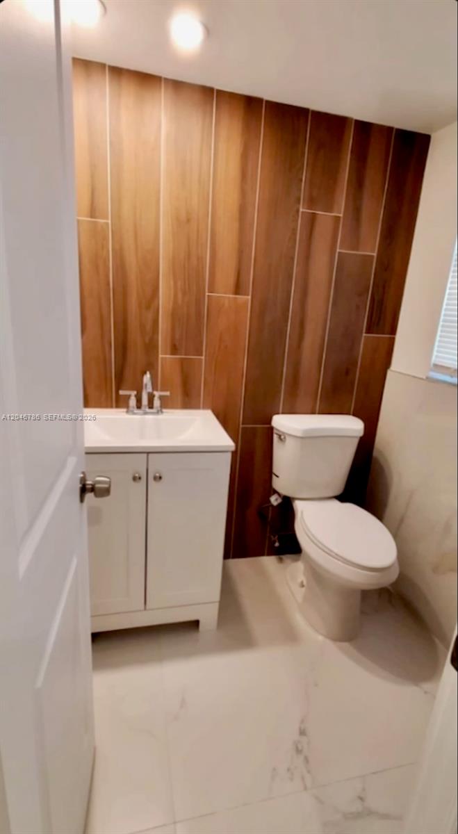 A small bathroom features a white vanity, a toilet, and a wood-look tile accent wall — 14790 Bonaire Blvd # 202, Delray Beach FL 33446, DELRAY BEACH, FL