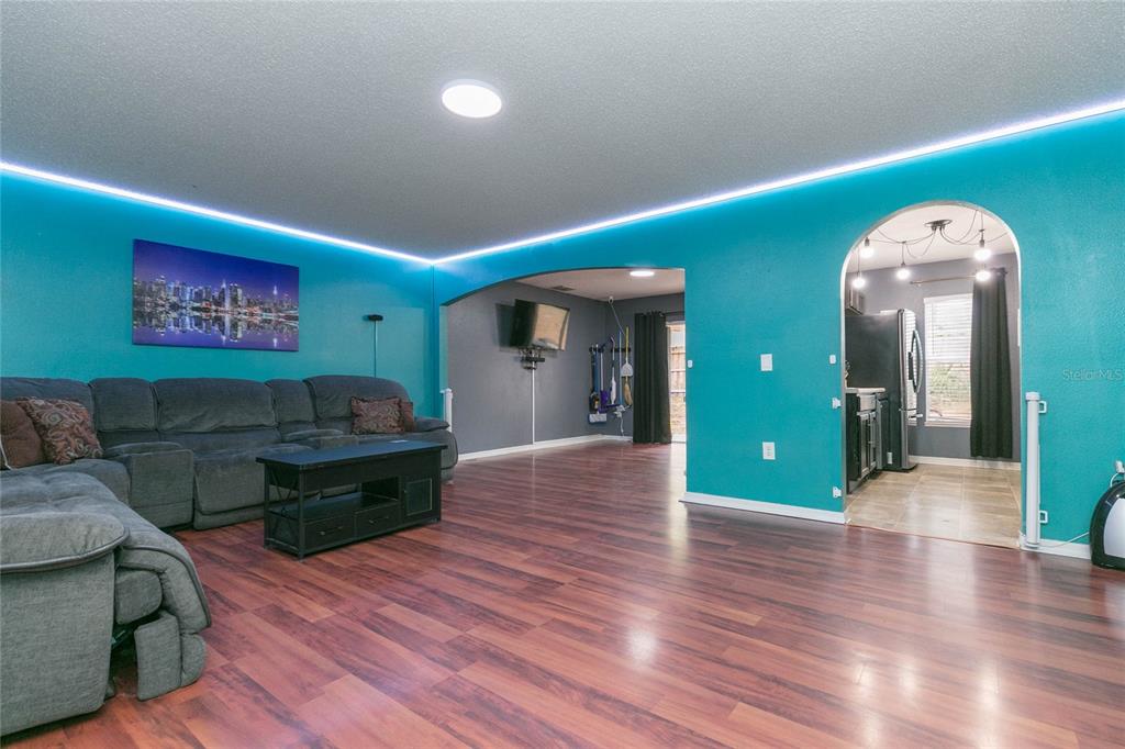 A living room with teal walls, LED cove lighting, and wood-look flooring leads to a kitchen — 9922 ASHBURN LAKE DRIVE, TAMPA, FL