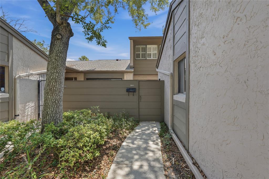 A concrete walkway leads to a gated entrance beside a stucco building and tree — 805 WESTWIND LANE, FERN PARK, FL