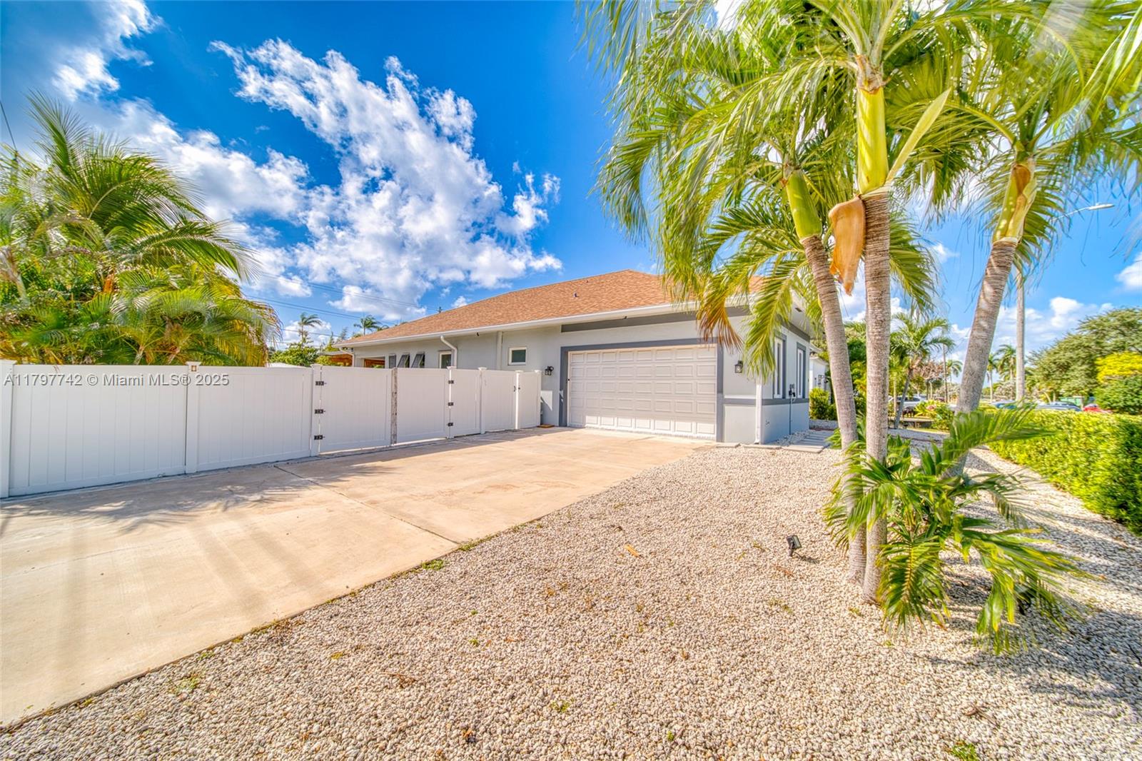 A single-story house with a white fence, concrete driveway, and gravel yard under a blue sky — 207 SW 11th St, Dania Beach FL 33004, DANIA BEACH, FL