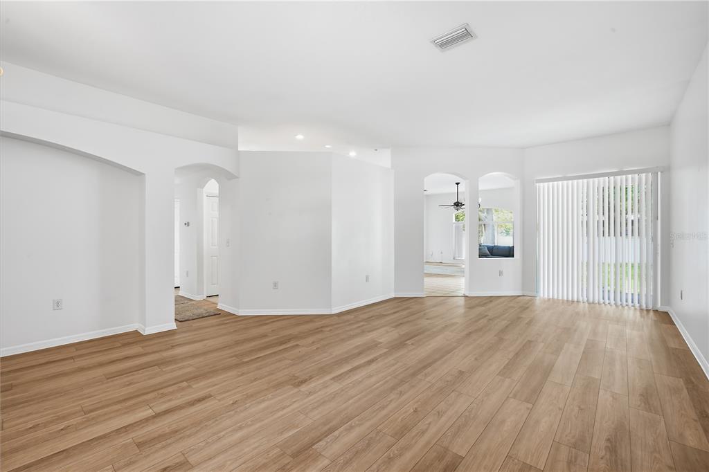 A spacious, empty living room with light wood-look flooring, white walls, and vertical window blinds — 5912 BRAMBLE BUSH COURT, ZEPHYRHILLS, FL