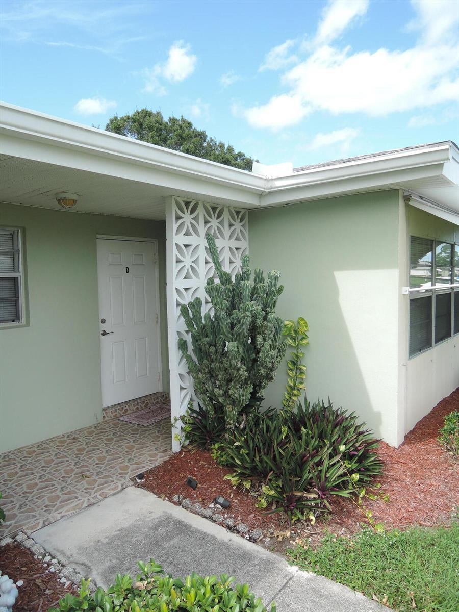 A light green exterior wall features a white door, a decorative breeze block screen, and landscaping — 115 Lakes End Drive Apt D, Fort Pierce FL 34982, FORT PIERCE, FL